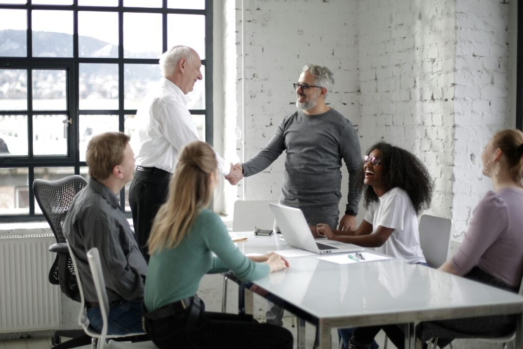 pexels-photo-3931440-3931440 Cheerful male colleagues shaking hands while discussing business ideas with group of multiethnic coworkers gathering around table with gadgets and documents in modern light workspace